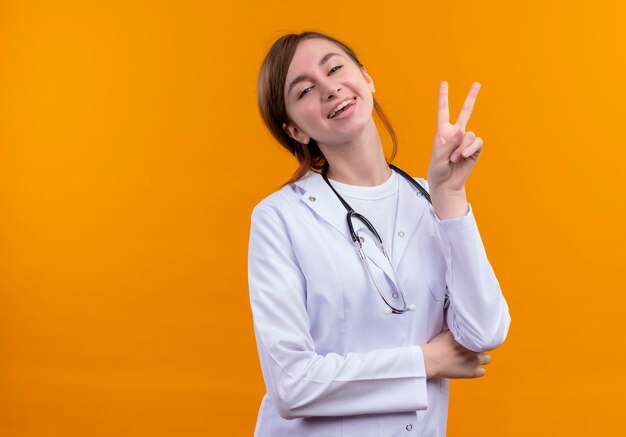 Alegre joven doctora vistiendo una bata médica y un estetoscopio haciendo el signo de la paz en el espacio naranja aislado con espacio de copia