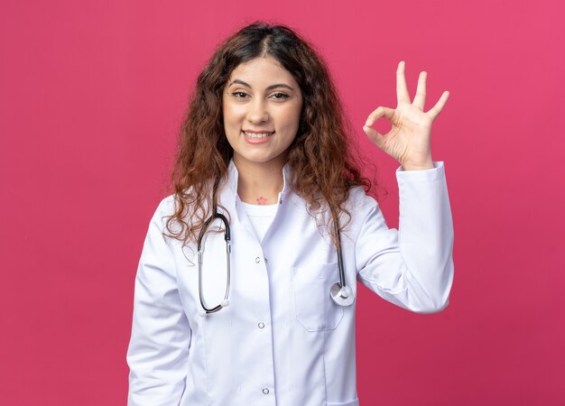 Alegre joven doctora vistiendo una bata médica y un estetoscopio mirando al frente haciendo el signo de ok aislado en la pared rosa