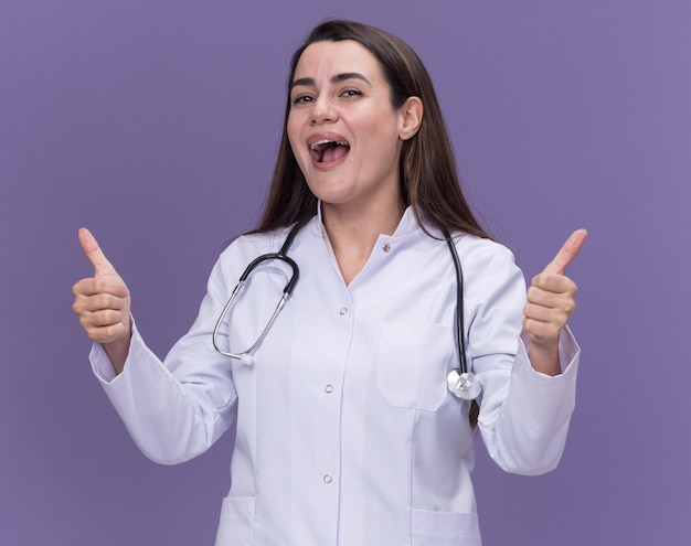 Alegre joven doctora vistiendo bata médica con estetoscopio pulgar hacia arriba con dos manos aisladas en la pared púrpura con espacio de copia