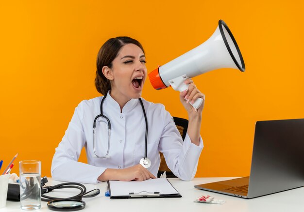 Alegre joven doctora vistiendo bata médica con estetoscopio sentado en el escritorio, trabajo en computadora con herramientas médicas, habla por altavoz en la pared amarilla de aislamiento