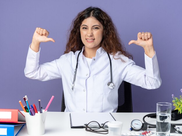 Alegre joven doctora vistiendo bata médica y un estetoscopio sentado en la mesa con herramientas médicas mirando al frente apuntando a sí misma aislada en la pared púrpura