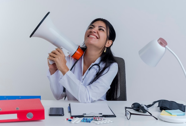 Alegre joven doctora vistiendo bata médica y un estetoscopio sentados en un escritorio con herramientas médicas sosteniendo el altavoz mirando al lado aislado