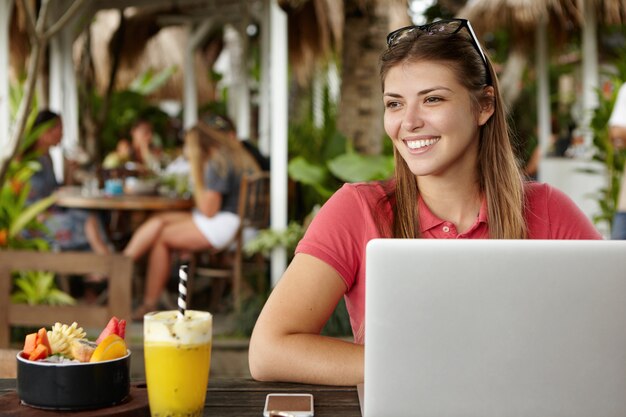 Alegre joven empresaria caucásica disfrutando días felices de sus vacaciones, sentada en la cafetería del hotel con dispositivos electrónicos y cócteles, revisando el correo electrónico en la computadora portátil