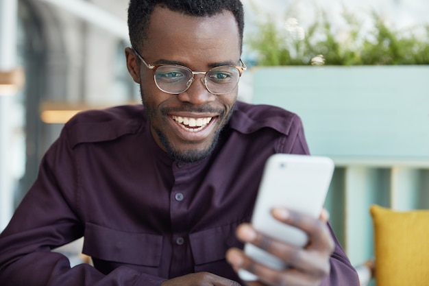 Alegre joven empresario con gafas redondas y ropa formal, verifica el suministro de noticias en un teléfono inteligente moderno, conectado a internet inalámbrico