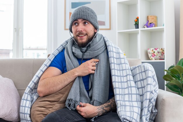 Alegre joven enfermo con bufanda y gorro de invierno con estetoscopio envuelto en una manta sentado en el sofá en la sala de estar mirando de lado escuchando sus propios latidos