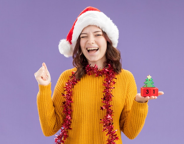 Alegre joven eslava con gorro de Papá Noel y con guirnalda alrededor del cuello parpadea y sostiene el adorno del árbol de Navidad aislado sobre fondo púrpura con espacio de copia