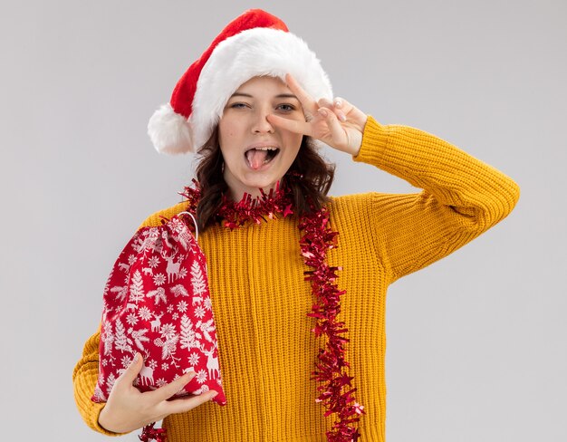 Alegre joven eslava con gorro de Papá Noel y con guirnalda alrededor del cuello saca la lengua y gestos signo de victoria con bolsa de regalo de Navidad aislado sobre fondo blanco con espacio de copia