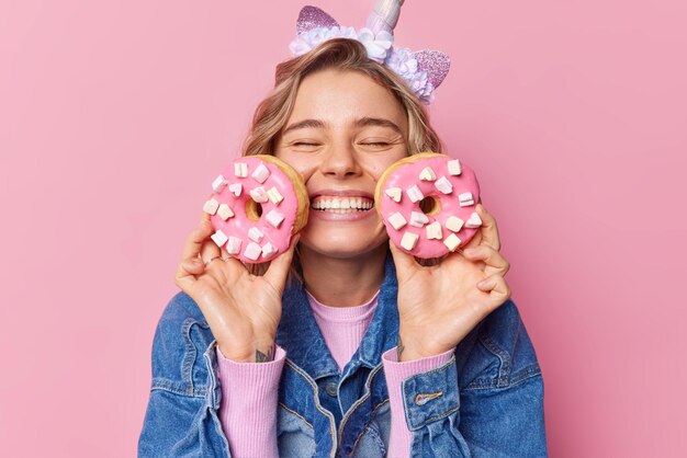 Alegre joven europea de cabello rubio mantiene los ojos cerrados sonrisas con dientes sostiene dos donuts con malvaviscos contentos de comer el postre favorito vestido con una chaqueta de mezclilla aislada sobre fondo rosa