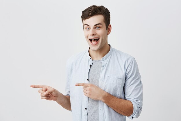 Alegre joven europeo con cabello oscuro y ojos azules, guiñando un ojo, vestido con una camisa azul que señala los dedos de distancia, indicando que hay espacio de copia en la pared blanca en blanco para contenido o información promocional