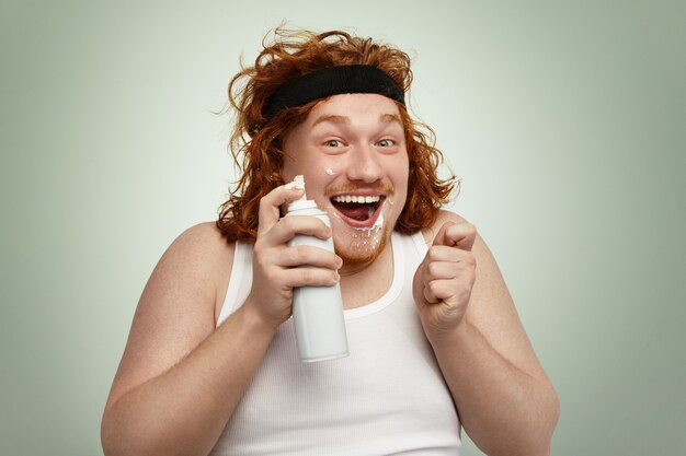 Alegre joven europeo gordito con cabello rizado de jengibre divirtiéndose en el interior, sosteniendo la lata de aerosol, su cara sucia con crema batida blanca, mirando con expresión facial feliz y emocionada