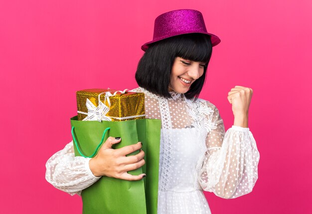 Alegre joven fiestera con gorro de fiesta sosteniendo el paquete de regalo en una bolsa de papel mirando hacia abajo haciendo un gesto de sí aislado en la pared rosa