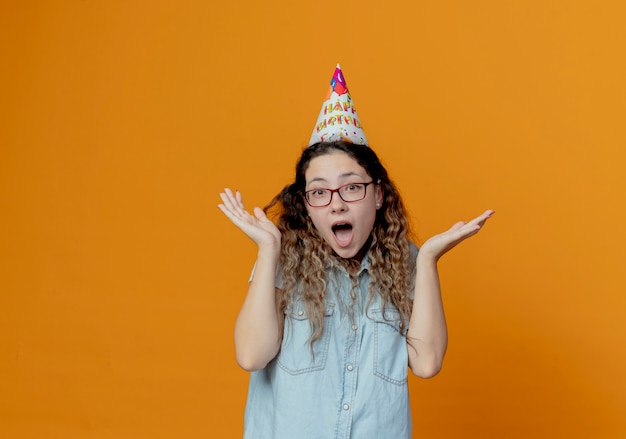 Alegre joven con gafas y gorro de cumpleaños extiende las manos aisladas sobre fondo naranja