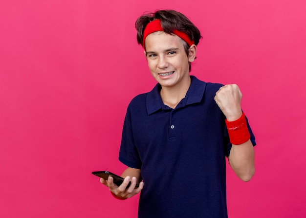 Alegre joven guapo deportivo con diadema y muñequeras con aparatos dentales que sostienen el teléfono móvil mirando al frente haciendo gesto de sí aislado en la pared rosa con espacio de copia