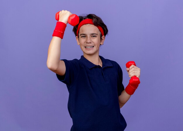 Alegre joven guapo deportivo vistiendo diadema y muñequeras con aparatos dentales levantando pesas aislado en la pared púrpura con espacio de copia