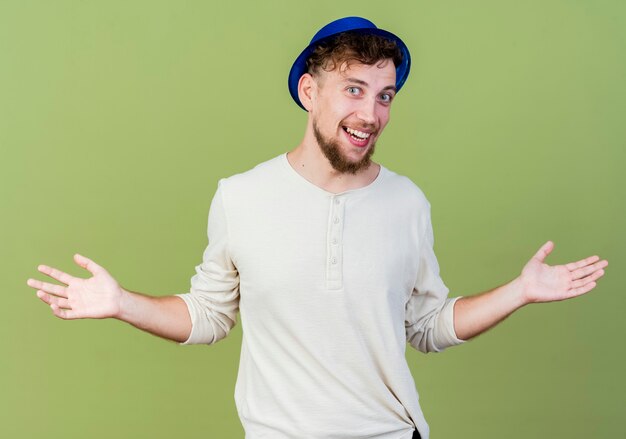 Alegre joven guapo partido eslavo con gorro de fiesta mirando a la cámara mostrando las manos vacías aisladas sobre fondo verde oliva