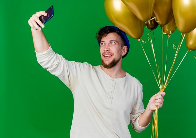 Alegre joven guapo partido eslavo con gorro de fiesta sosteniendo globos tomando selfie aislado sobre fondo verde