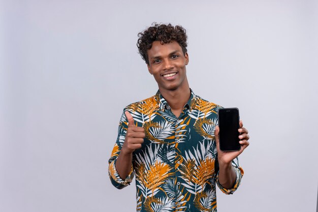 Alegre joven guapo de piel oscura con cabello rizado en hojas camisa estampada que muestra una pantalla de teléfono inteligente en blanco con los pulgares hacia arriba