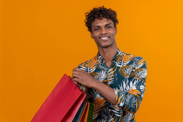 Alegre joven guapo de piel oscura con cabello rizado en hojas camisa estampada sonriendo sosteniendo bolsas de compras mientras está de pie sobre un fondo naranja