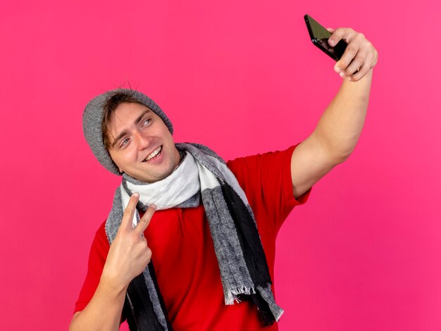 Alegre joven guapo rubio enfermo vestido con gorro y bufanda de invierno haciendo el signo de la paz tomando selfie aislado en la pared carmesí