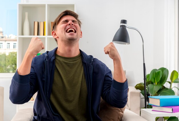 Alegre joven guapo rubio se sienta en un sillón manteniendo los puños y mirando hacia arriba