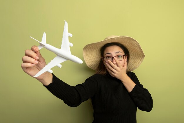 Alegre joven hermosa en un cuello alto negro y gafas sosteniendo un avión de juguete conmocionado cubriendo la boca con la mano