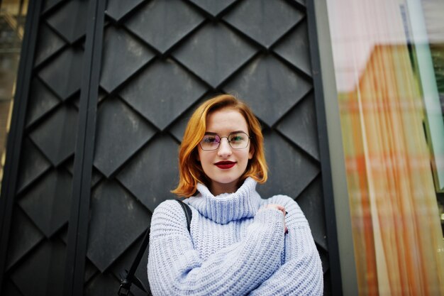 Alegre joven hermosa mujer pelirroja en gafas cálido suéter de lana azul con mochila posada al aire libre