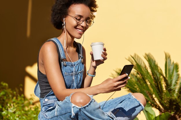 Alegre joven hipster con cabello rizado y tupido, usa anteojos y overoles andrajosos, descarga canciones a la lista de reproducción del teléfono móvil, bebe bebidas frescas de un vaso desechable, disfruta del tiempo libre durante el verano