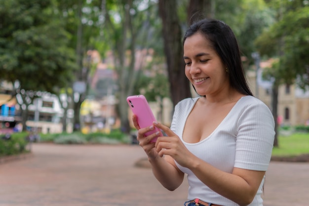 Alegre joven hispana sentada en un parque y enviando mensajes de texto por teléfono