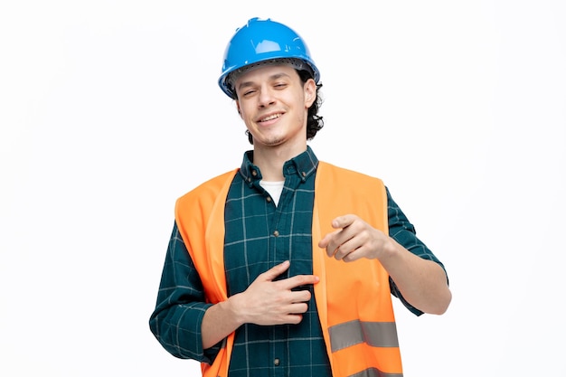 Alegre joven ingeniero masculino usando casco de seguridad y chaleco de seguridad manteniendo la mano en el vientre mirando y apuntando a la cámara riéndose aislada en el fondo blanco