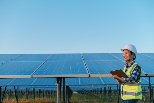 Alegre joven inspectora asiática ingeniera tocando para verificar la operación del panel solar fotovoltaico en la estación al aire libre sosteniendo el tablero de la lista de verificación con espacio de copia de sonrisa