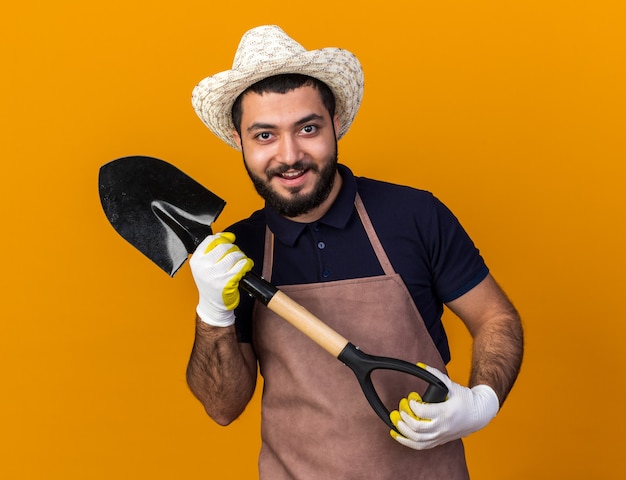 Alegre joven jardinero varón caucásico vistiendo gorro de jardinería y guantes sosteniendo la pala aislado en la pared naranja con espacio de copia