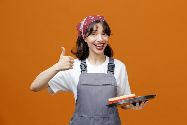 Alegre joven limpiadora vistiendo uniforme y bandana sosteniendo una bandeja con una esponja mirando a la cámara mostrando el pulgar hacia arriba aislado en el fondo naranja