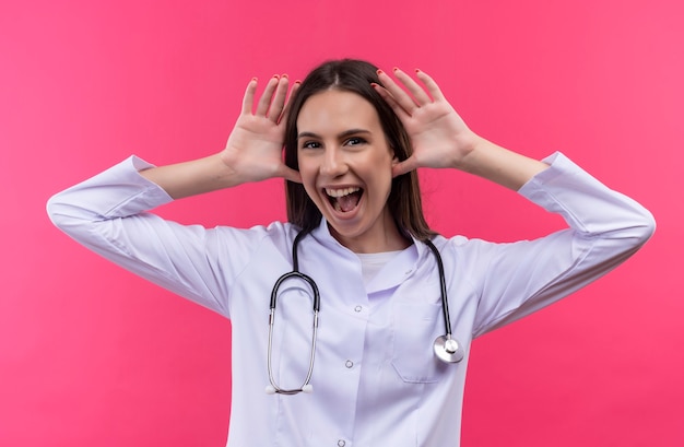 Alegre joven médico con bata médica estetoscopio puso sus manos alrededor de las orejas en la pared rosa aislada