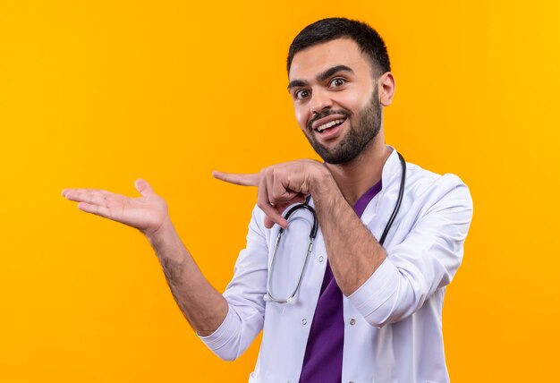 Alegre joven médico con estetoscopio bata médica apunta al lado con el dedo en la pared amarilla aislada
