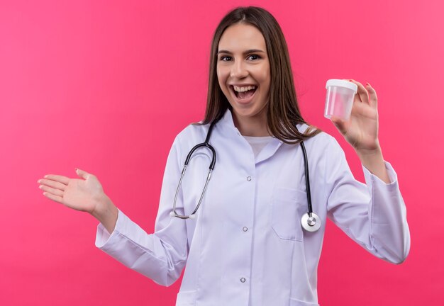 Alegre joven médico con estetoscopio bata médica sosteniendo lata vacía en la pared rosa aislada