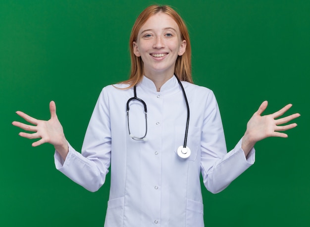 Alegre joven médico de jengibre vistiendo bata médica y un estetoscopio mostrando las manos vacías aisladas en la pared verde