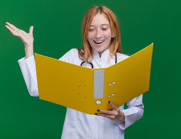 Alegre joven médico de jengibre vistiendo bata médica y un estetoscopio sosteniendo y mirando la carpeta que muestra la mano vacía aislada en la pared verde