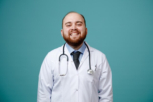 Alegre joven médico masculino con abrigo médico y estetoscopio alrededor del cuello mirando a la cámara sonriendo aislado en el fondo azul