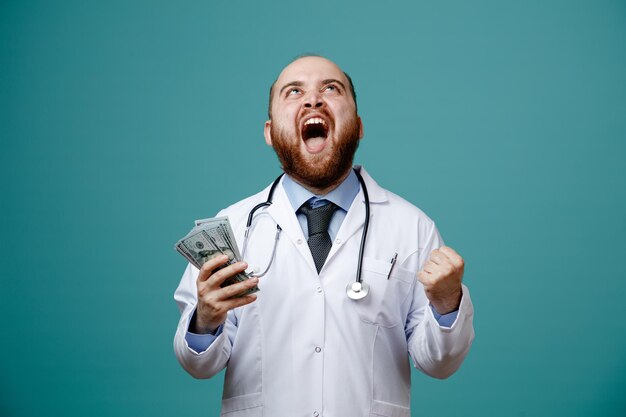 Alegre joven médico masculino con abrigo médico y estetoscopio alrededor del cuello sosteniendo dinero mirando hacia arriba mostrando un gesto de sí aislado en el fondo azul