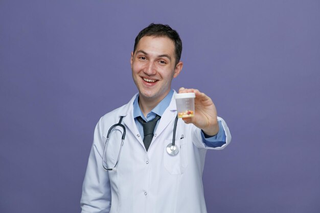 Alegre joven médico masculino con bata médica y estetoscopio alrededor del cuello mirando a la cámara estirando el contenedor de medidas hacia la cámara aislada en el fondo morado