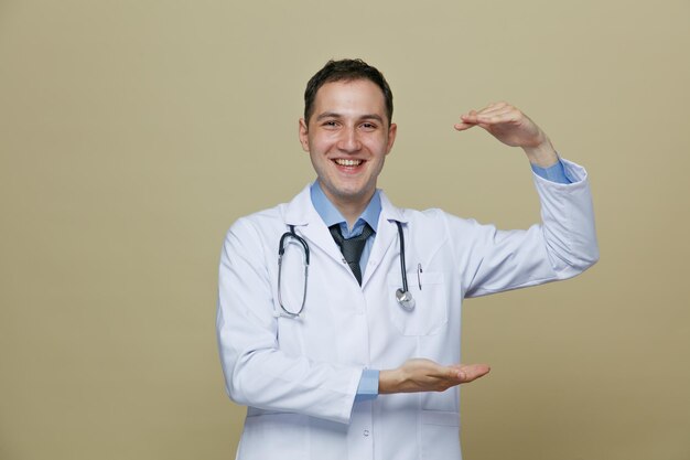 Alegre joven médico masculino con bata médica y estetoscopio alrededor del cuello mirando a la cámara mostrando un gesto de tamaño aislado en un fondo verde oliva
