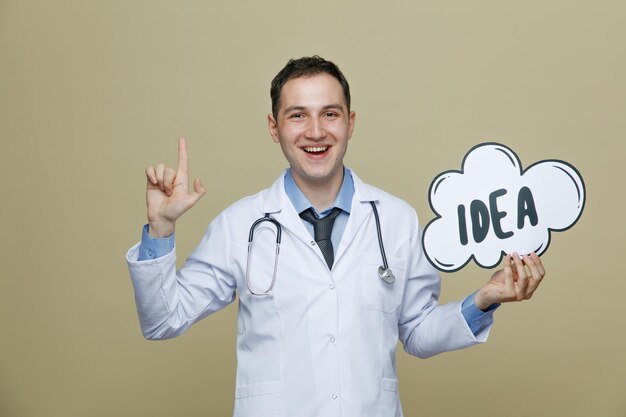 Alegre joven médico masculino con bata médica y estetoscopio alrededor del cuello mostrando una burbuja de ideas mirando a la cámara apuntando hacia arriba aislada en un fondo verde oliva