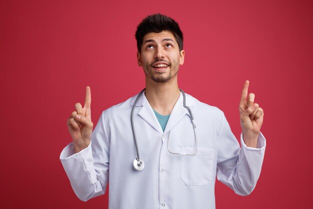 Alegre joven médico masculino con uniforme médico y estetoscopio alrededor del cuello mirando hacia arriba señalando con el dedo aislado en el fondo rojo