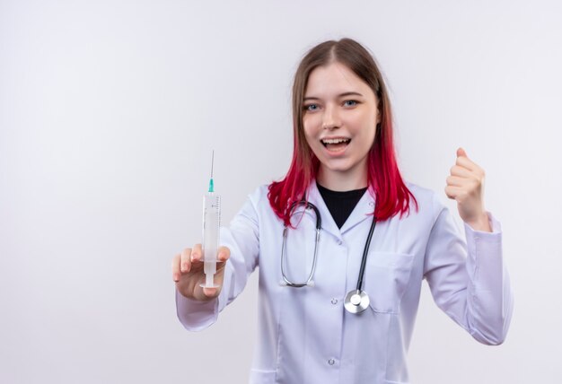 Alegre joven médico mujer vistiendo estetoscopio bata médica sosteniendo una jeringa levantando el puño en la pared blanca aislada