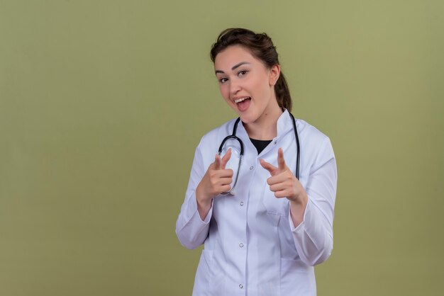 Alegre joven médico vistiendo bata médica con estetoscopio apunta hacia adelante en la pared verde