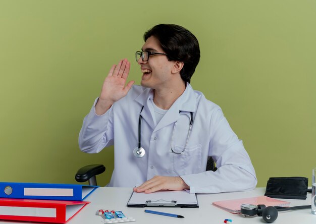 Alegre joven médico vistiendo bata médica y un estetoscopio con gafas sentados frente al escritorio