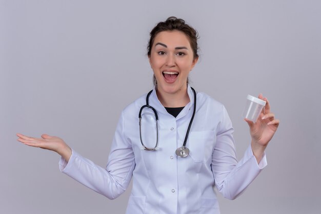 Alegre joven médico vistiendo bata médica con estetoscopio sosteniendo lata vacía en la pared blanca