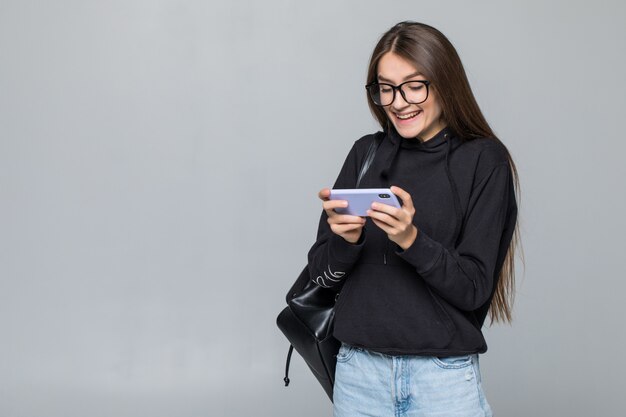 Alegre joven con mochila juego con teléfono móvil aislado en la pared blanca.