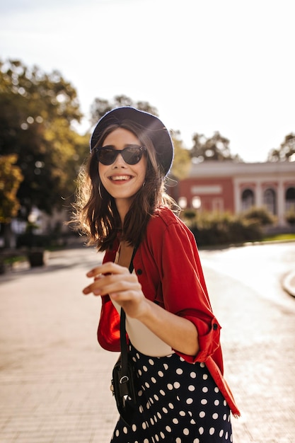 Alegre joven morena con camisa roja superior blanca de boina y gafas de sol sonriendo y posando al aire libre Ciudad soleada de verano y clima cálido