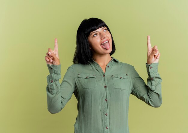 Alegre joven morena caucásica saca la lengua y apunta hacia arriba con las dos manos aisladas sobre fondo verde oliva con espacio de copia